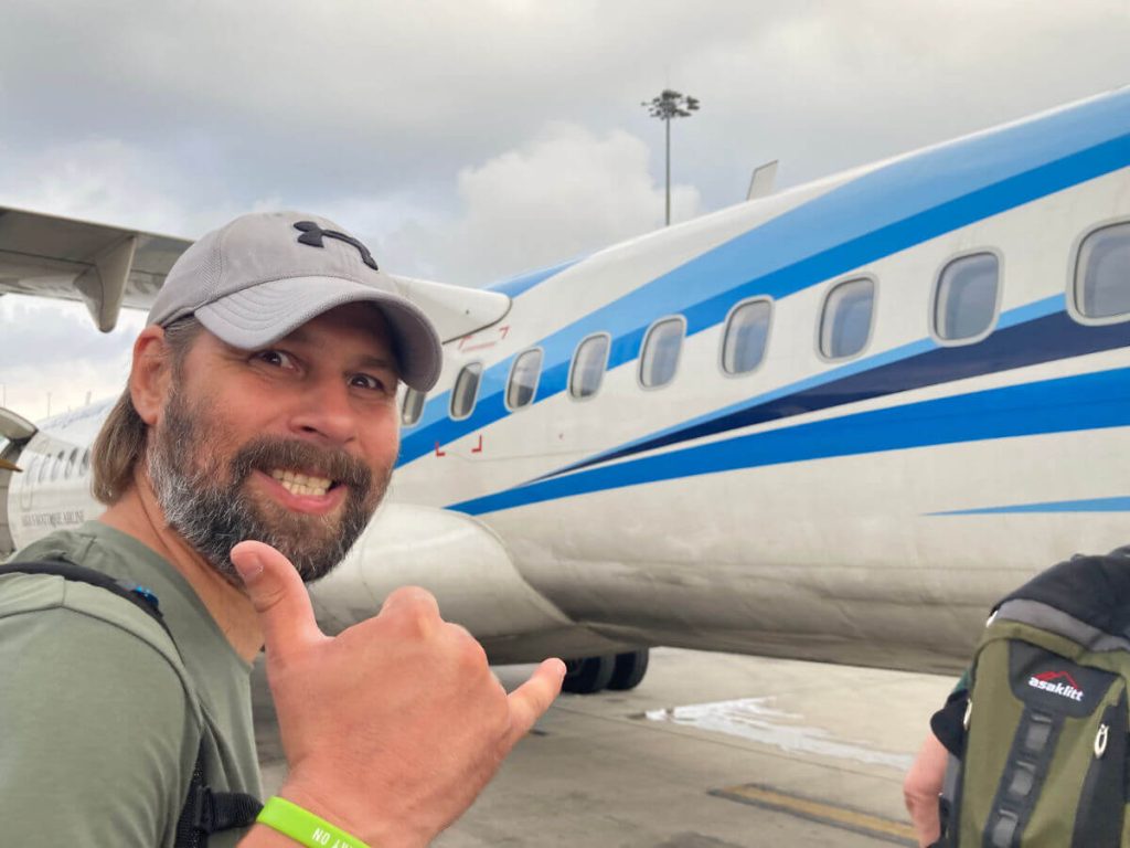 Man in a green t-shirt and grey baseball cap making a 'cool' sign with fingers in-front of a blue propeller plane