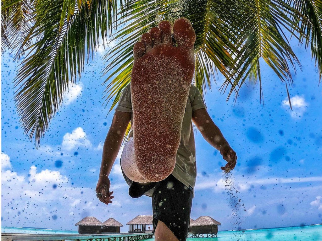 The bottom of a mans right foot held up in front of the camera covered in sand on a beach
