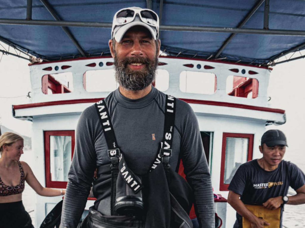 A Man in a grey hat and white sunglasses standing on the deck of a boat with a SANTI drysuit wrapped around his waist