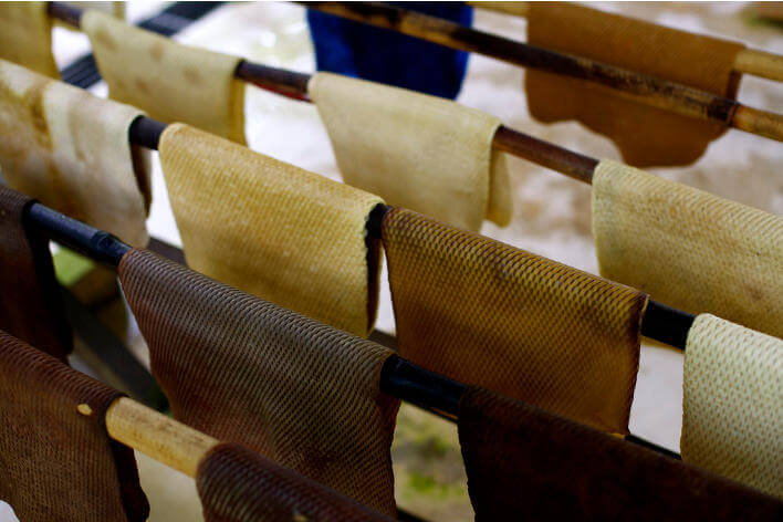 Sheets of natural rubber in various shades of black, grey and cream hanging over wooden poles