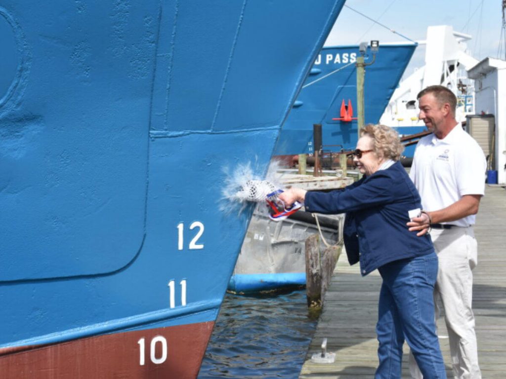 The man helping a woman smash a bottle of champagne onto the bow of a large blue boat