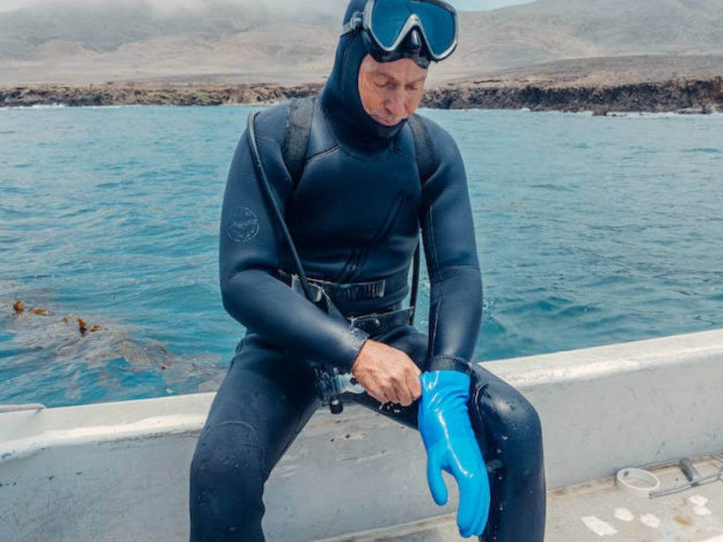A man sitting on the edge of a boat in a semi-drysuit putting on a blue rubber glove