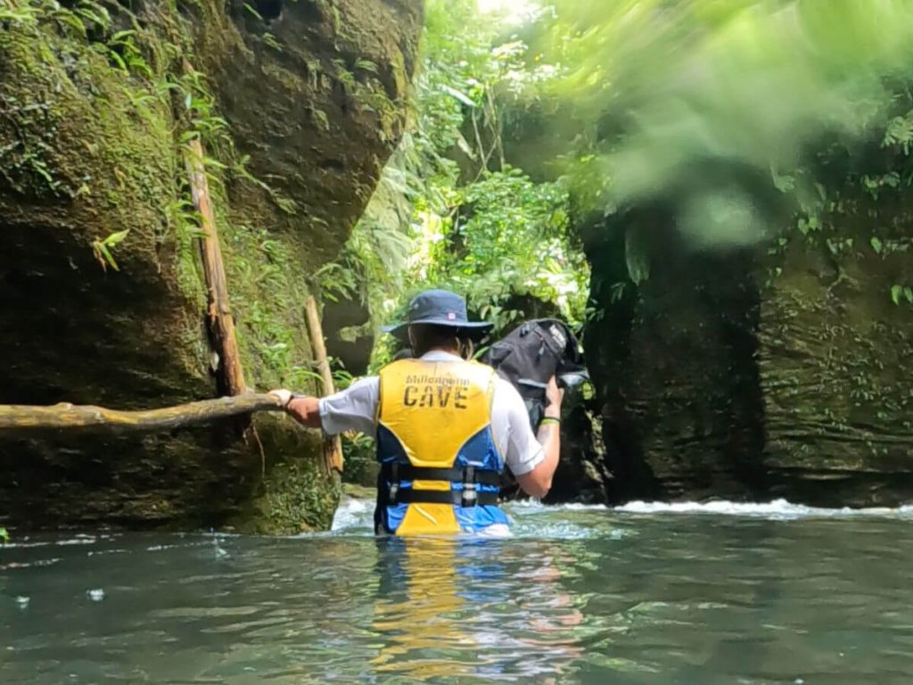 Man wading through waist deep water holding a small black backpack in the air